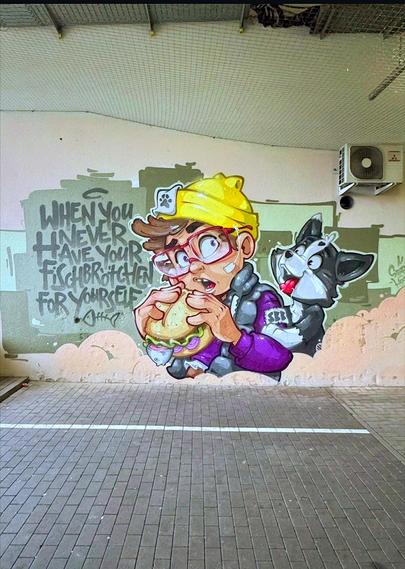 Streetartwall. A funny comic mural of a boy with a penchant for fish sandwiches and cats is sprayed on a wall in Berlin. The figure with short brown hair under a yellow cap and purple jacket is standing there and is about to bite into one of these fish sandwiches. Behind him, on his arm, sits a cheerful black and white cat, whose favorite food also seems to be these fish Sandwiches. The figure looks open-mouthed through his new red glasses (the purchase label is still on) and notices that the cat is trying to get a bite. Next to it is the text: "When you never have your Fischbrötchen for yourself."