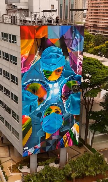 Streetartwall. A beautiful, huge, six-storey mural with four doctors was sprayed/painted on the outside of a public hospital. The mural is very colorful and designed as a large-scale mosaic in the typical style of the artist "Kobra". It shows four faces (male and female) with face masks and surgical clothing looking down on an imaginary patient from above. The lamps of the operating theater shine through between them from above in the middle. The faces of the figures are painted in bright, shining colors, only the surgical clothing is blue. It is intended to honor the people in this hospital who save lives through their careful, precise and compassionate work.
Note: The photo shows the artist on the roof of the building.The building is also a place of art. It was idealized by Oscar Niemeyer, the gardens are by Burle Marx and inside there is a mosaic by Di Cavalcanti.