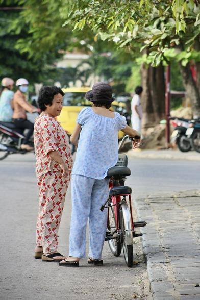 das bild im hochformat zeigt zwei frauen beim smalltalk an einer straßenecke in einer vietnamesischen stadt.
beide frauen tragen zweiteilige bekleidung, vielleicht pyjamas. eines ist hellblau gemustert, das andere rötlich.
während die linke frau von der seite zu sehen ist – sie lächelt und hört ihrer gesprächspartnerin zu –, ist die andere frau nur von hinten zu sehen. sie hat ein rotes fahrrad bei sich.
im hintergrund sind behelmte motorradfahrer_innen, ein parkendes auto und bäume zu sehen.