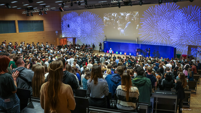 Foto von einem vollen Hörsaal bei der Feierlichen Immatrikualtion. Das Publikum steht und schaut in Richtung eines Sprechers.