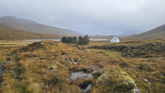 A remote white building stands out against the muted browns and greys of the Scottish Highlands. Rain falls softly, blurring the outlines of the distant mountains. The lake in the valley reflects the somber hues of the sky. Despite the dreary weather, the landscape exudes a sense of quiet beauty.  The vastness of the scenery evokes a feeling of solitude and peace.