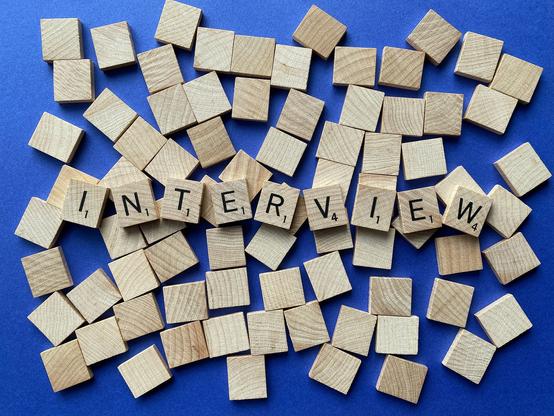 Das Wort "Interview" aus Scrabble-Steinen gelegt. Im Hintergrund lauter umgedrehte Scrabble-Stein auf blauem Grund.