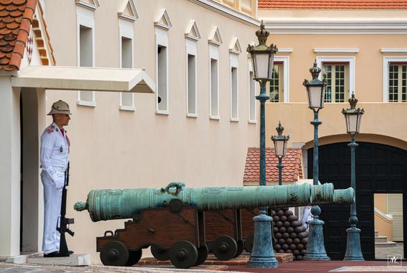 Color photo of a white-clad palace guard at a sentry station in front of Le Palais Princier de Monaco. In front of the guard are a row of three old cannons and a pile of cannon balls.