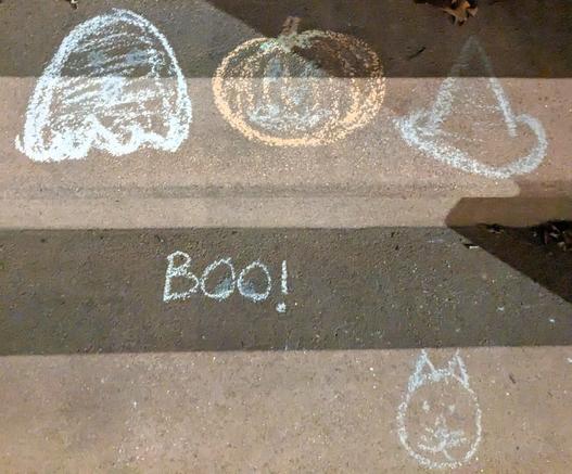 Chalk art seen on two steps on a brownstone stoop in Cobble Hill, Brooklyn. On the top step from left to right, we have a Pacman-esque ghost, a pumpkin, and a witch's hat. On the side of the top step, we see "Boo!" written out. On the bottom right of the next step down, there is a cat's face.