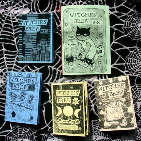 Photo of five small displayed on a black cloth with a white spider web design. The zines have cute DIY drawings of a cat on a guitar broom, tarot cards, the moon in various phases, and a shoulder bag for witchcraft on the go.