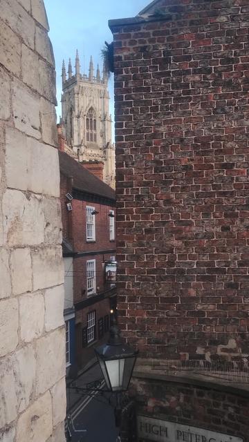 Glimpse of a Gothic tower of York Minster between old brick buildings and edge of a medieval city wall gatehouse