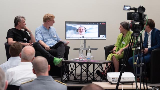 Das Bild zeigt die Paneldiskussion beim HSK-Endphasensystem der Cyberagentur mit fünf Personen. Vier sitzen in Sesseln um einen runden Tisch, auf dem Wasserflaschen und Gläser stehen. Eine fünfte Person ist auf einem Monitor im Hintergrund zu sehen und nimmt virtuell an der Diskussion teil. Im Vordergrund ist eine Kamera zu erkennen, die die Veranstaltung aufzeichnet. Die Teilnehmenden halten Mikrofone, während das Publikum aufmerksam zuhört. 
Foto: Cyberagentur