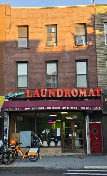 Photograph of a shop front with large glass or plastic red ketters spelling LAUNDROMAT and green script spelling 'super. The shop awning saus 355 smith street