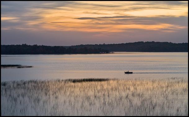 The sunset is partially hidden behind scattered clouds and there is a silouette of a boat on a calm lake with scattered swamp grass in the forground.