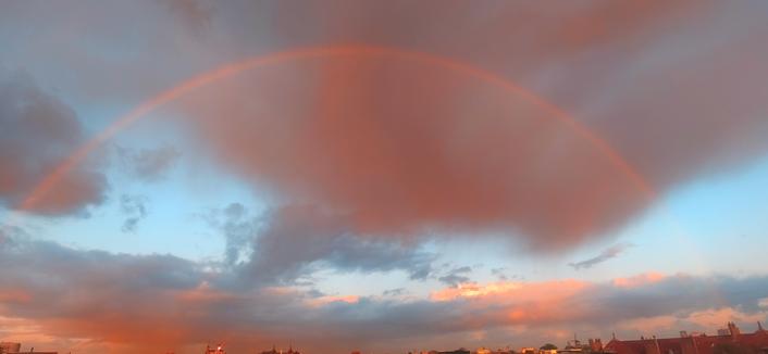 Sky view of grey pink rainclouds between a blue sky catching the red and yellow glow of sunrise. The raindrops show a beautiful half rainbow over the city of Amsterdam