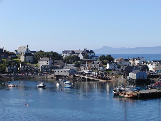 Mallaig. The image shows a view from a high point down across water onto a harbour that stretches across the centre of the frame and has moored boats, a slipway and boatyard and wharves with more boats. There are two and three storey houses and flats beyond the harbour and in the background on the right is sea that extends out to a distant island. The sky is blue.