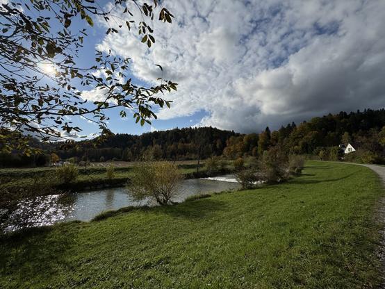 Panoramic view of autumn colored trees at the end of the valley. The upper right half is dominated by scattered clouds with a dark cloud base. The sun is shining in the left upper half. A river floats through the middle of the photo.