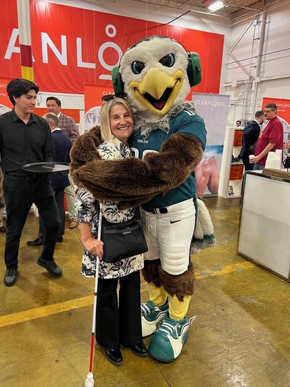 One woman, holding a white cane, stands smiling with the Philadelphia Eagle's team mascot, Swoop, an eagle in a green jersey. The scene is in a large indoor venue with several people in the background.
