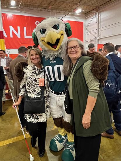 Two women stand smiling with the Philadelphia Eagle's team mascot, Swoop, an eagle in a green jersey. The scene is in a large indoor venue with several people in the background. One woman holds a white cane.