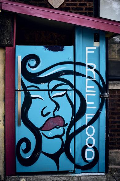 This image features a bright blue fridge with a mural painted on it. The mural depicts a woman's face with closed eyes and a relaxed expression, embodying a sense of calm and welcoming. Large, bold letters spell out 'FREE FOOD' next to the face, making it clear that the fridge provides free food for those in need.