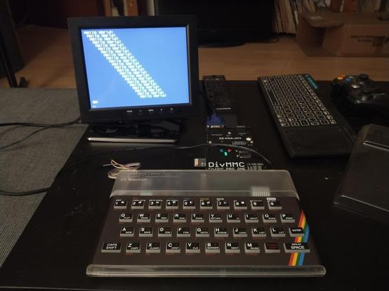 A Sinclair ZX Spectrum 128K clone (Harlequin 128K)  in a transparent, classic "rubber key" case with anodized titanium faceplate. It's connected to a 7" LCD displaying the text "Hello World!" 22 times, each line shifting the text by one space.