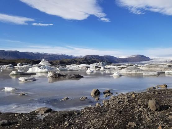 Icebergs illuminated by the sun in a mostly frozen lake. The shore in front is mostly gravel and a few boulders. A mountain range and glacier are visible in the background.