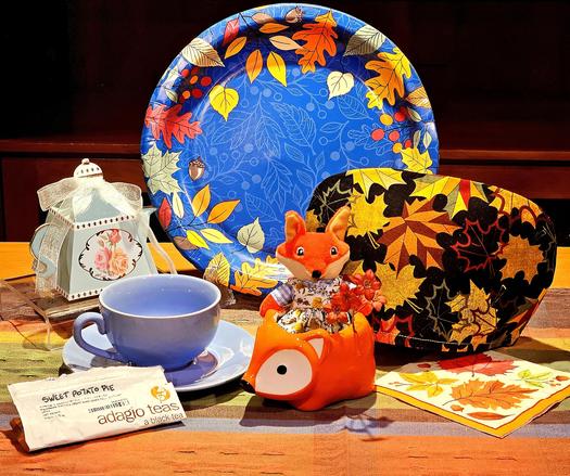 A tea tableau in Autumn colors punctuated with bright blue, like a crisp Fall day. Against a background design of Fall leaves and a leafy tea cozy, there is a fox doll in a country dress, holding a bouquet of orange flowers and sitting in a fox-shaped vessel or vehicle. The tea is Sweet Potato Pie black tea from Adagio.