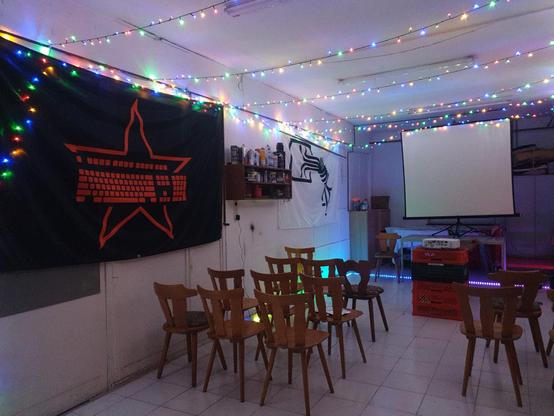 Zu sehen ist einer gefliester (Werkstatt)raum mit Stühlen, einem Beamer mit Leinwand, Lichterketten und Neopixel an Decke und Boden und Chaosflaggen an der Wand