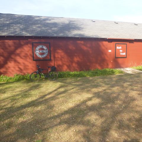 A view of a old rail terminal building along the Cannon Valley Trail with two large signs.  One is a recreation of the old railroad sign for the original Chicago Great Western Railroad.  The symbol is a circular design with the railroad identification along the outside of the circle.  Across the middle of the sign is a railroad marketing identification as the Corn Belt Route.  The second sign is a square with “Rail to Trail” as words arranged diagonally from top left to bottom right.  The building is a long, low single story structure painted a dark red with a light gray roof.