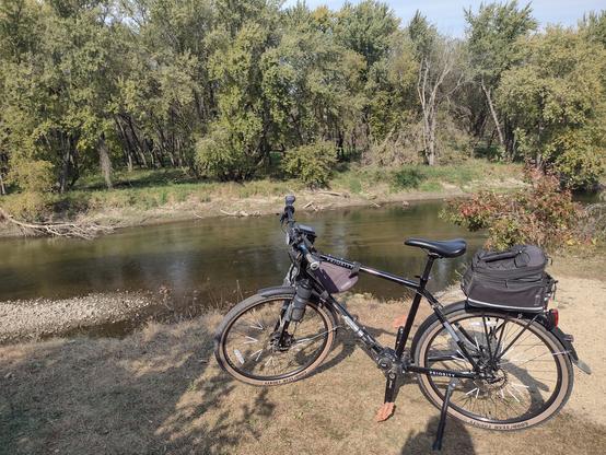 A view of a black bicycle posed along a trail on a river bank above the river.  The river is behind the bicycle from left to right.  On the far bank of the river green grass and mature trees are visible.