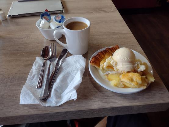 A image of a piece of apple pie with ice cream on a plate.  A mug of coffee and table utensils are to the left of the plate on the table.