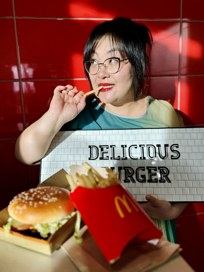 The author of this post sits in front of a red wall eating a french fry while holding an awkward looking sign that says DELICIOUS BURGER. In front of her is a burger and fries with a McDonald’s logo.