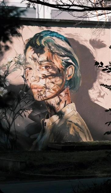 Streetartwall. A beautiful portrait of a woman was sprayed/painted on the outside wall of a modern one-story building. It shows a young woman with short (green) hair standing between fine branches with white flowers. Her face is somewhat obscured because the branches cast shadows on her face and on the beige wall behind her. This wonderful portrait, which looks like a fine Asian drawing, lives from this sophisticated play of shadows. A homage to Henri Matisse.
Info: Henri Matisse's quote "There are flowers everywhere for those who want to see them" invites us to take a new perspective on the world around us. It expresses a philosophy of life in which the beauty and richness of nature are accessible to those who have the sensitivity to perceive them.