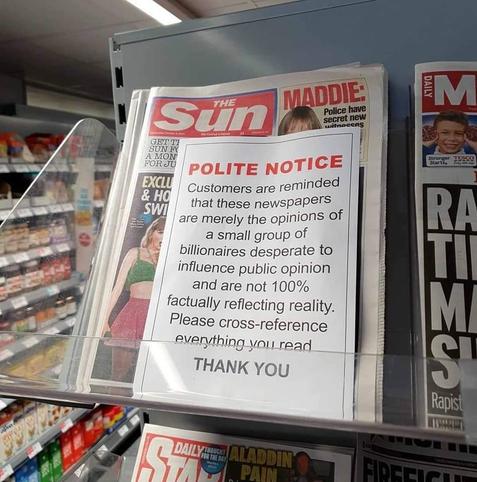 A display in a store featuring a tabloid newspaper with a prominent notice warning customers that the content is influenced by wealthy individuals and may not reflect reality. The message advises readers to cross-reference information.