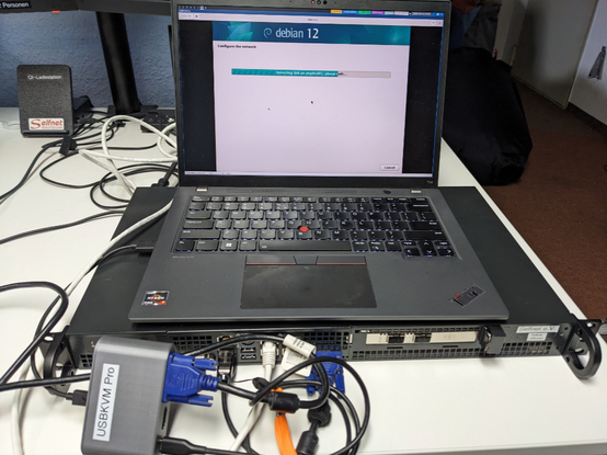 A small 1U server with a laptop on top. The  laptop shows the Debian installer. It's connected to the server with a USBKVM Pro.