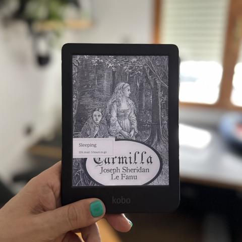 Mano sujetando un lector de libros electrónicos con la portada de Carmilla: como un grabado a una tinta aparecen dos mujeres jóvenes con un bosque de fondo.