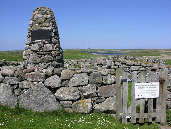 Flora MacDonald's birthplace and monument. The image shows an enclosure made of large stones in the foreground enclosing a circular cairn with a plaque on it. There’s a gate in the bottom right of the frame with a welcome sign on it. The landscape in the background is very flat. The sky is blue.
