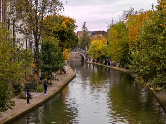 A serene canal scene with people walking along tree-lined paths, framed by autumn foliage. A stone bridge arches in the distance, adding depth to the cozy atmosphere. The water reflects the trees and overcast sky, enhancing the tranquil setting.