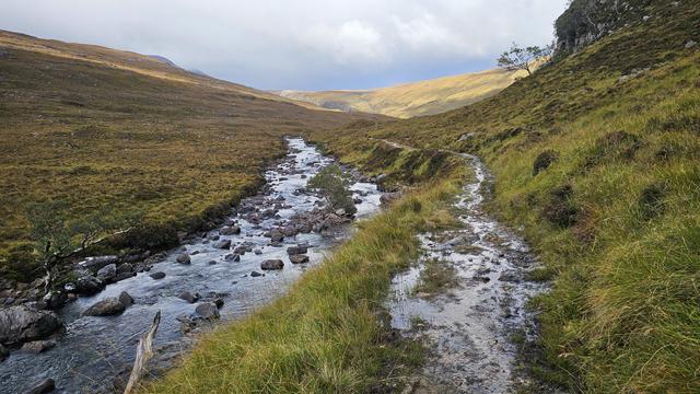 Scenic landscape of a tranquil stream flowing through a lush, grassy valley under a cloudy sky. The path beside the river is completely flooded with water.