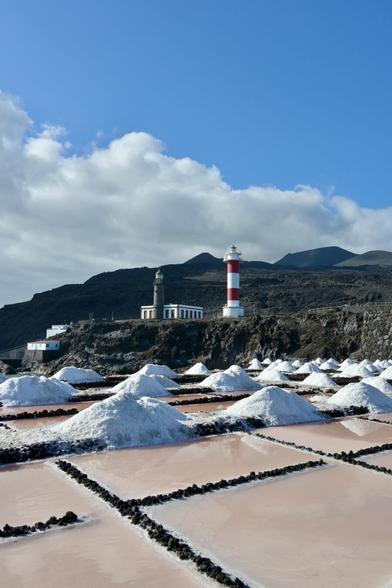Two lighthouses in the background, sitting beneath dark volcanic peaks, with pinkish salt pans in the foreground.