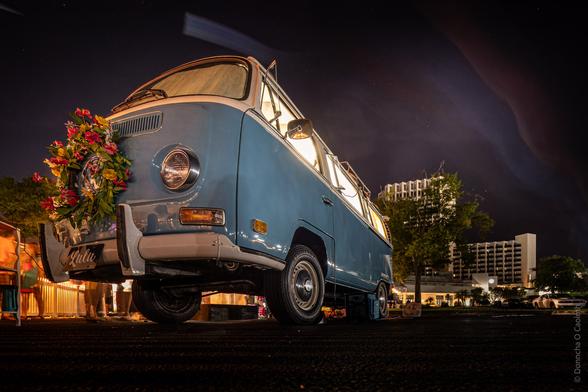 A vintage blue Volkswagen van adorned with a colourful flower wreath on its front. The van is parked outdoors at night, with city buildings visible in the background. The scene is illuminated by warm lights, giving it a festive and lively atmosphere. The license plate reads "Lulu."