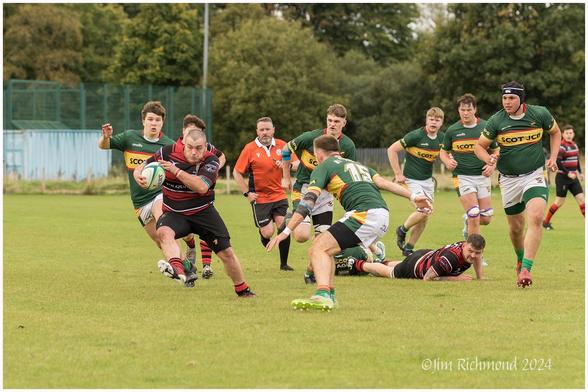 A rugby match is taking place on a grassy field. One player, wearing a red and black jersey, is running with the ball while being pursued by opponents in green jerseys. Several players are engaged in the play, with some on the ground.