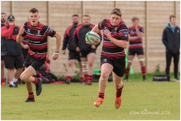A rugby player runs on the field, carrying a rugby ball, while another player follows closely behind. In the background, teammates watch from the sidelines. The players wear red and black striped jerseys.