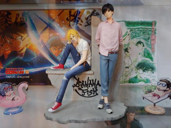 Figure of Ash and Eiji from Banana Fish. Ash has blond hair, a white shirt, and a blue jeans and sit on a low wall, hand and head resting on his knee. He's peeking at Eiji, who leaning against the wall, hands behind his back. Eiji wears blue pants, a pale pink shirt, and has dark hair