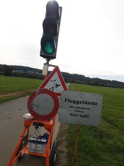 Ein asphaltierter Feldweg, auf dem eine mobile Ampel mit einem "Durchfahrt verboten"-Schild, einem Hinweisschild mit der Aufschrift "Fluggelände - Bei erhobener Fahne hier halt!", sowie einem Warnschild mit dem Piktogramm eines Fußgängers, der durch die Schleppleine an einem Segelflugzeug halbiert wird.