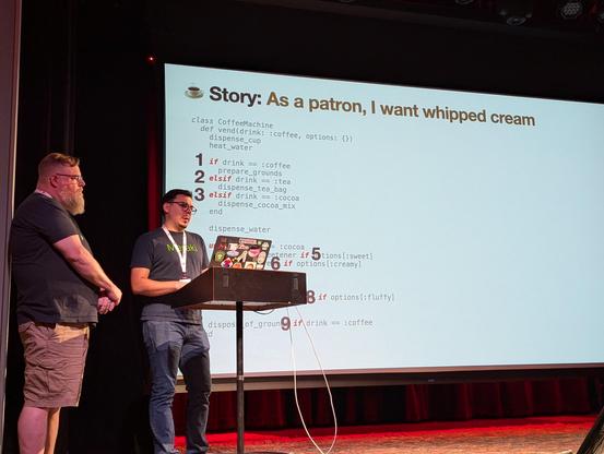 Fito von Zastrow (with Alan Ridlehoover) speaking to the Rocky Mountain Ruby audience in front of the a slide from a talk called The Coffee Machine Talk: A Brewer's Guide to Filtering out Complexity and Churn. The slide says "Story: As a patron, I want whipped cream" and includes Ruby code with the nine conditionals highlighted.