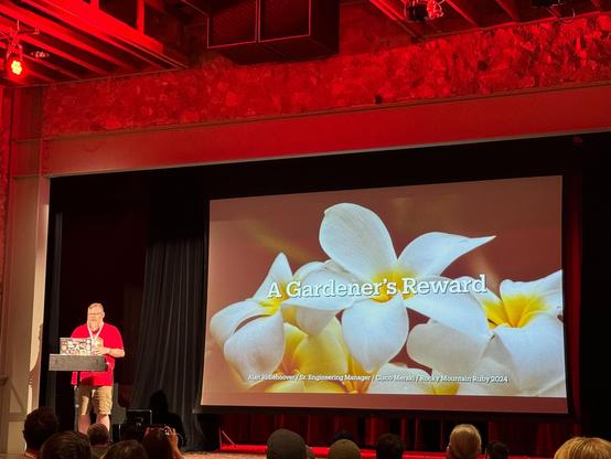 Alan Ridlehoover speaking to the Rocky Mountain Ruby audience in from of the title slide of a presentation entitled: "A Gardener's Reward." The title slide has a picture of plumeria flowers behind the title text.
