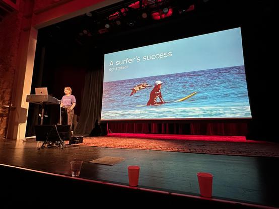 Liz Heym speaking to the Rocky Mountain Ruby audience behind a lecture and in front of a slide with a photograph of Liz on a surf board in calm waters with a pelican flying by her right shoulder. The title of the slide is "A surfer's success."