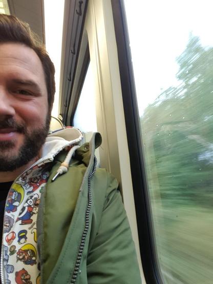 Das Bild ist ein Selfie eines Mannes, der sich gerade in einem fahrenden Zug befindet und halb zu sehen ist. Die andere Hälfte. Die Person freut sich und trägt unter anderem einen Super-Mario-Hoodie.
