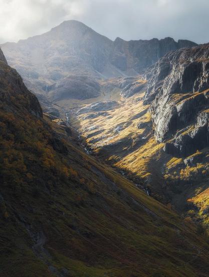 A breathtaking view of Corrie nan Lochan nestled between two of the Three Sisters of Glencoe, showcasing the raw, rugged beauty of the Scottish Highlands. The image captures a dramatic mountain landscape bathed in golden autumnal light. Steep, craggy slopes rise on either side of a narrow glen, their rocky faces etched with shadows. A winding stream cascades down the centre, glinting in the sunlight. The vegetation on the hillsides ranges from rich golden hues to deep browns, indicating the changing seasons. In the background, a majestic mountain peak looms, partially obscured by mist, adding to the ethereal atmosphere. The interplay of light and shadow accentuates the contours of the landscape, creating a sense of depth and grandeur that epitomises the wild, untamed nature of Glencoe.