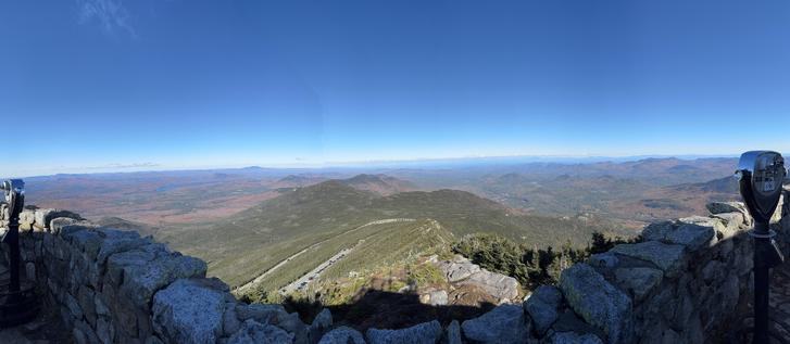 The view to the north (towards Montréal):

We can see mountains left, right and centre. The trees are green and orange hues. There are no clouds in the sky.
There is a road (NY-431) coming up the mountain with a long queue of cars. 

At the nearest point from the photographer, there is a stone wall to prevent people from accidentally falling. On the immediate left and right, there are two lookout thingees.
- - -
Vue vers le nord (direction de Montréal):

On peut voir des montagnes dans toutes les directions. Les arbres sont dans des teintes de vert et d’orange. Il n’y a pas de nuage dans le ciel.
Il y a une route qui monte la montagne (NY-431) avec une longue file d’attente.

Au point le plus proche du photographe, il y a un mur de pierre pour éviter que les gens tombent accidentellement. Sur la gauche et la droite immédiate, il y a des trucs d’observation.