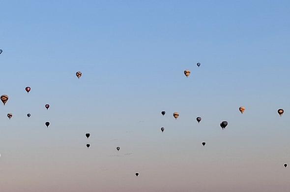 A hazy morning sky that is light blue near the top and gradually shows a light, reddish gray band at the bottom. Scattered throughout the sky are tiny dots, which are actually hot air balloons. Some of the balloons show bands and colors of red, orange, and yellow while others are too dark to display any distinguishing colors.