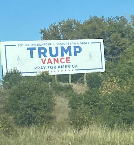 A picture of a white billboard with blue and red lettering in southern Wisconsin showing "secure the border" mispelled as "boarder". It is a Trump-Vance "pray for America" billboard with two images of praying hands.  Across the top it says "Secure the boarder [sic]" and "Restore law and order".