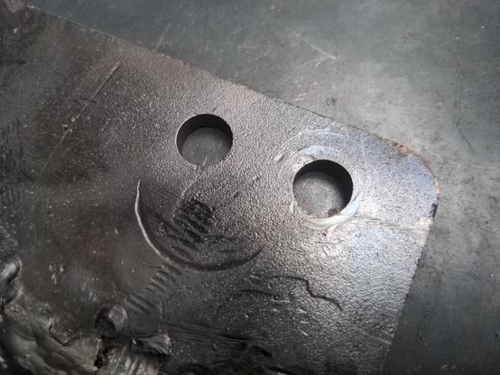 A close-up of a piece of 6mm thick steel with two holes in it. Below the holes is a semi-circular indentation, caused by the force used on it in a hydraulic press. It makes it look like a happy face.