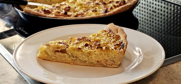 Ein Stück Zwiebelkuchen auf einem Teller vor einem runden Blech mit mehr Zwiebelkuchen.
